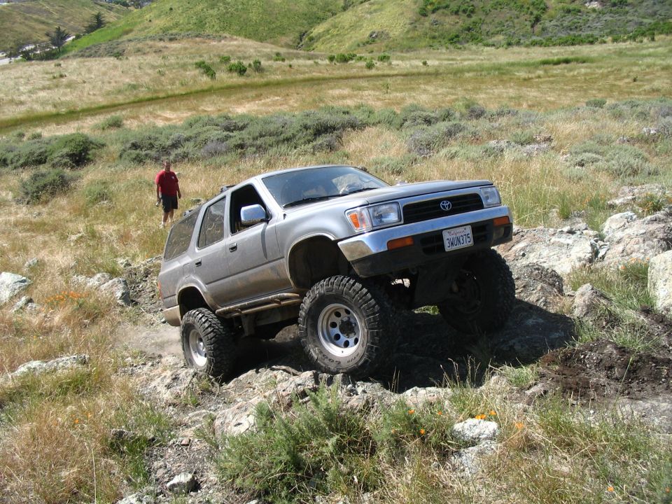 Silver lifted Toyota 4Runner flexing over rocks on a hillside, person walking in background