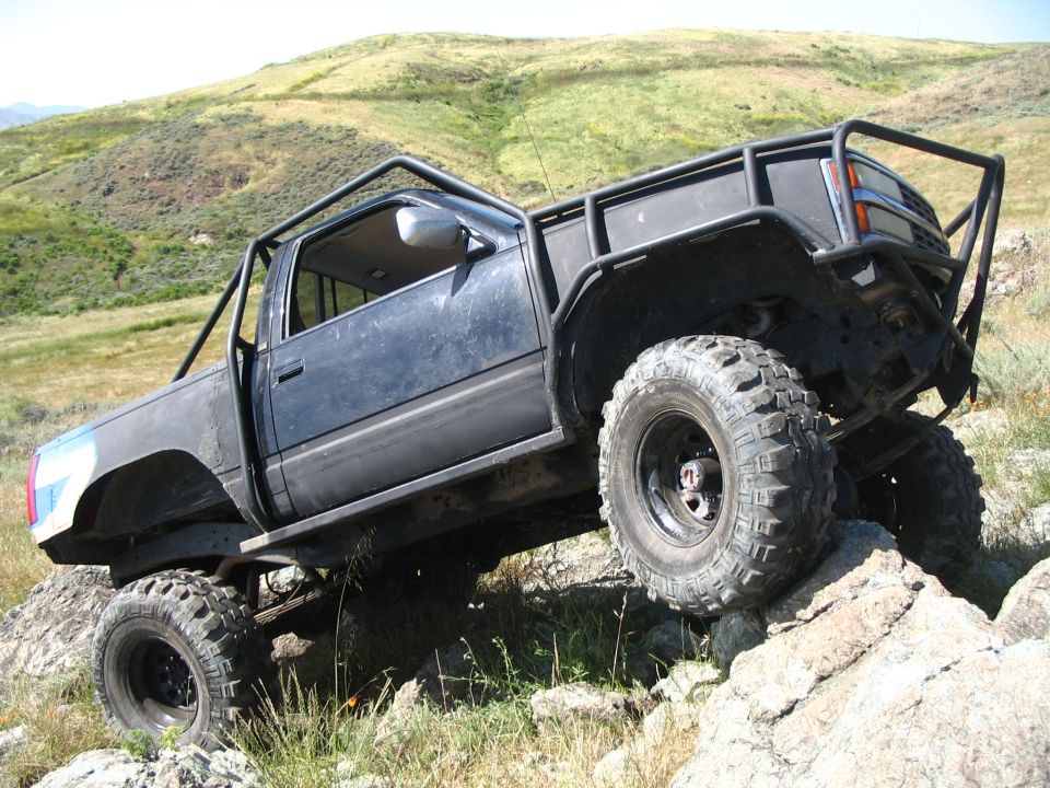 Black tube-bodied lifted mini truck on huge tires perched on rocks above a green valley