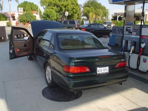 1995 Ford Taurus - A green 1995 Ford Taurus sedan is parked at a gas station with its hood and driver's door open.