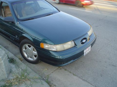 1995 Ford Taurus - A green 1995 Ford Taurus is parked by the curb, with a red car passing by in the background.