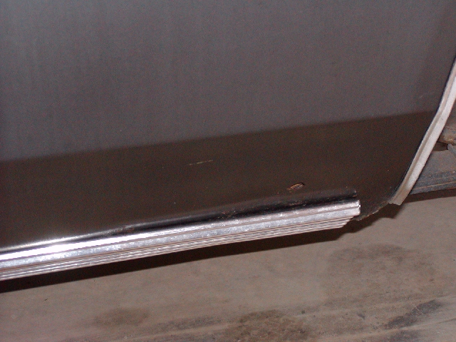 Close-up of a dark-colored vehicle's lower body panel with a chrome trim piece. Some rust is visible