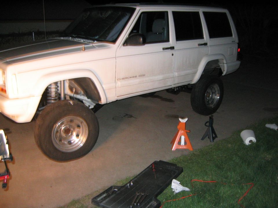 White Jeep Cherokee with lifted suspension, front wheel off, on jack stands. Tools and oil drain pan nearby