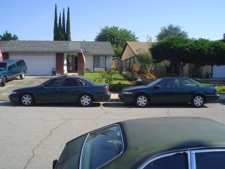 Two dark green 1995 Ford Taurus sedans parked side-by-side on a street in front of houses