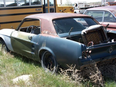1968 Ford Coupe - A weathered Ford Mustang coupe sits in a junkyard, showing signs of significant disrepair and missing components.