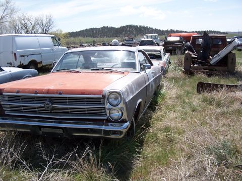 1968 Ford Coupe - A weathered 1966 Ford sits in a junkyard with other vehicles visible in the background under a cloudy sky.