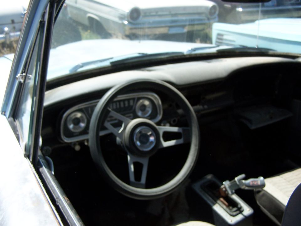 Interior of a 1968 Ford Coupe showing dashboard, steering wheel, and gear shift. Another car is visible through the windshield