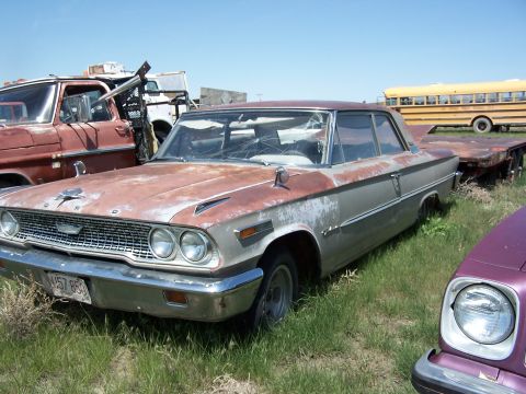 1968 Ford Coupe - A weathered 1963 Ford Galaxie sits in a junkyard next to a tow truck and a yellow school bus.