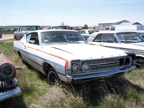 1968 Ford Coupe - A white 1968 Ford Coupe with red racing stripes sits in a junkyard, surrounded by other vehicles.
