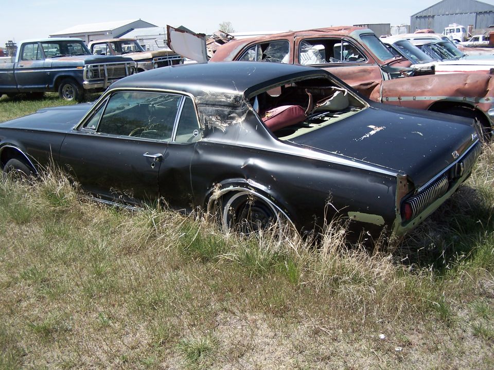 Abandoned black 1967 Mercury Cougar coupe in a junkyard, overgrown with grass. Other vehicles and buildings are in the background