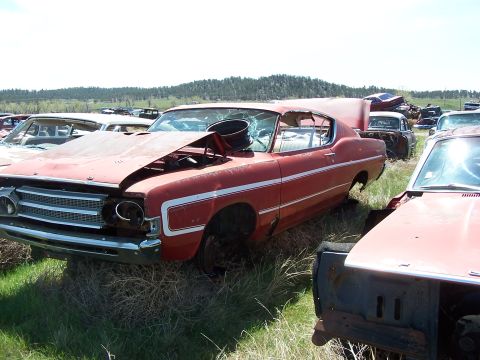 1968 Ford Coupe - This red coupe sits in a junkyard, showing signs of significant wear and damage, including a broken windshield and missing parts.