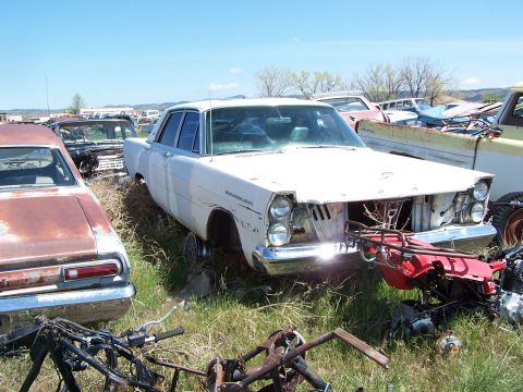 1968 Ford Coupe - A white sedan sits in a junkyard, surrounded by other vehicles and debris under a clear blue sky.