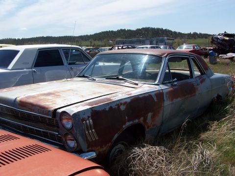1968 Ford Coupe - A heavily rusted 1966 Ford Fairlane coupe sits in a junkyard, surrounded by overgrown grass and other vehicles.