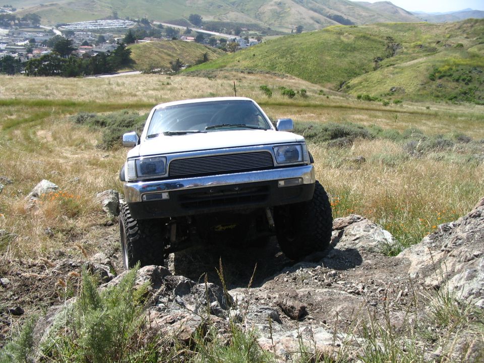 White lifted Toyota with billet grille climbing rocks on a coastal hill, town in valley