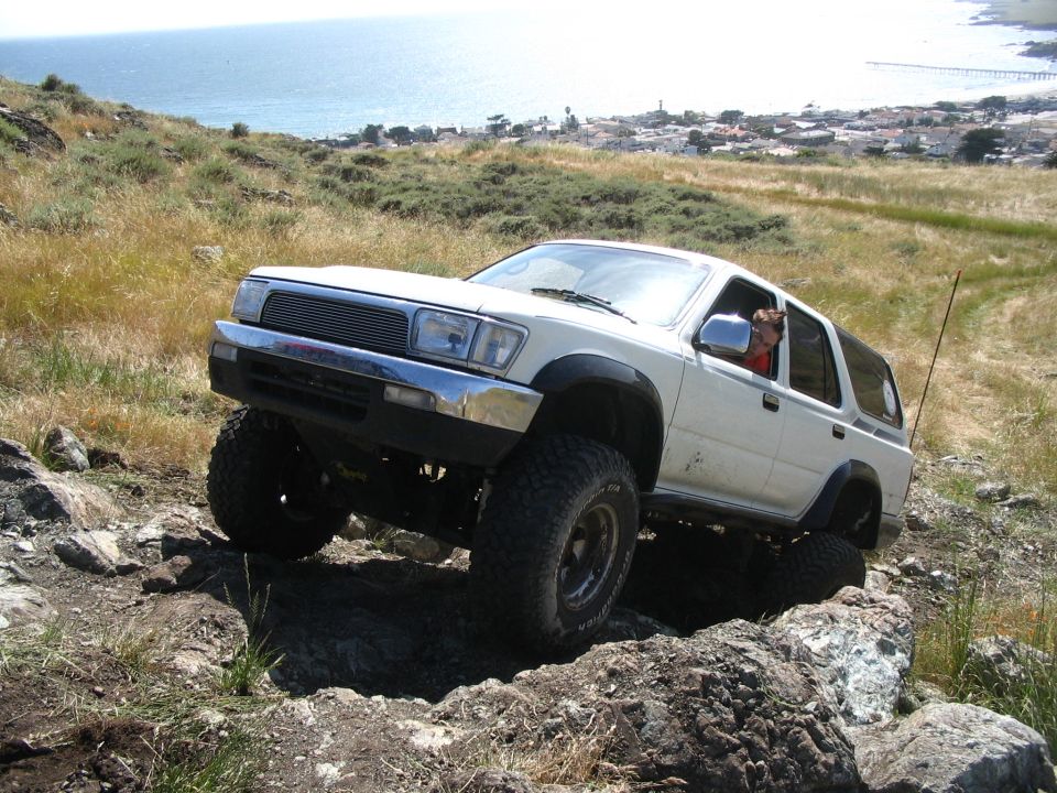 White lifted Toyota truck flexing over rocks on a hill above an ocean coastline town