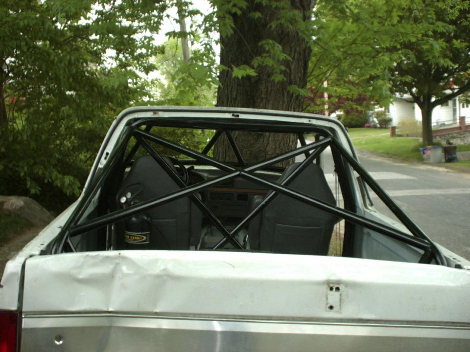 White pickup truck's interior with a black roll cage, two bucket seats, and a fire extinguisher. The rear of the truck is dented