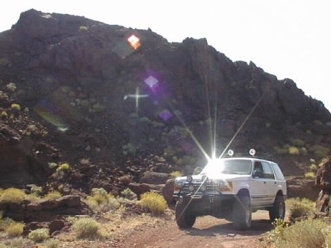 1997 Ford Ranger - A white SUV equipped with auxiliary lights navigates a dusty trail amidst a , mountainous terrain.