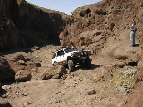 1997 Ford Ranger - A white SUV is driving through a rocky canyon. A person is standing on the cliff above watching.