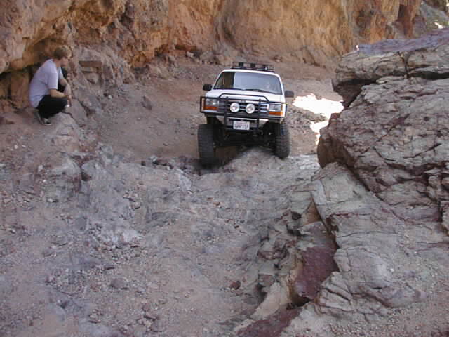 White off-road vehicle driving on a rocky trail in a canyon. Person crouches on the left side of the trail