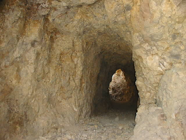 Mine tunnel with rough rock walls and a bright opening in the distance. Supermotors.net logo in the corner