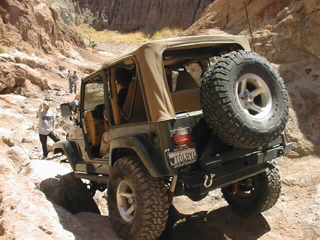 Black Jeep Wrangler with tan soft top and spare tire on rear, driving on rocky terrain