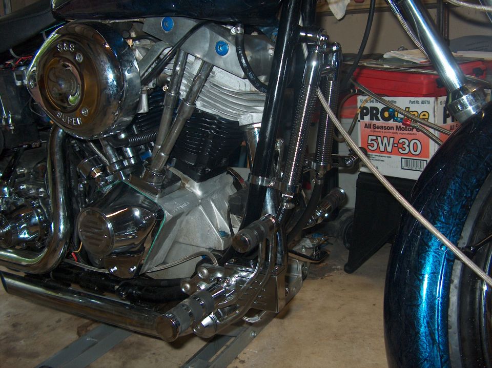 Motorcycle engine with S&S Super air cleaner, chrome exhaust pipes, and foot controls. Oil container visible in background