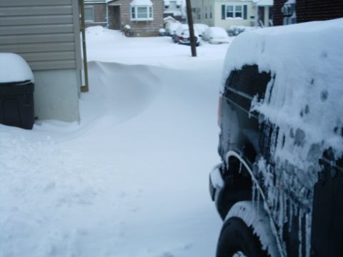1989 Ford Bronco - A black SUV is partially visible, covered in snow and icicles, with other snow-covered vehicles and houses in the background.