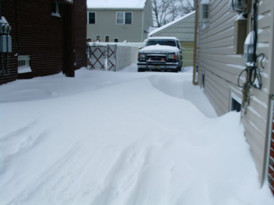 Snow-covered Ford Bronco parked in a snowed-in driveway between two houses after a heavy storm