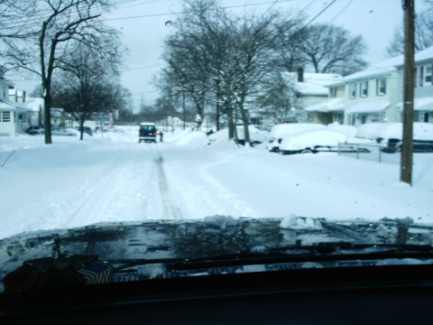 1989 Ford Bronco - A dark SUV navigates a snow-covered residential street, with parked cars buried in snow on the sides.