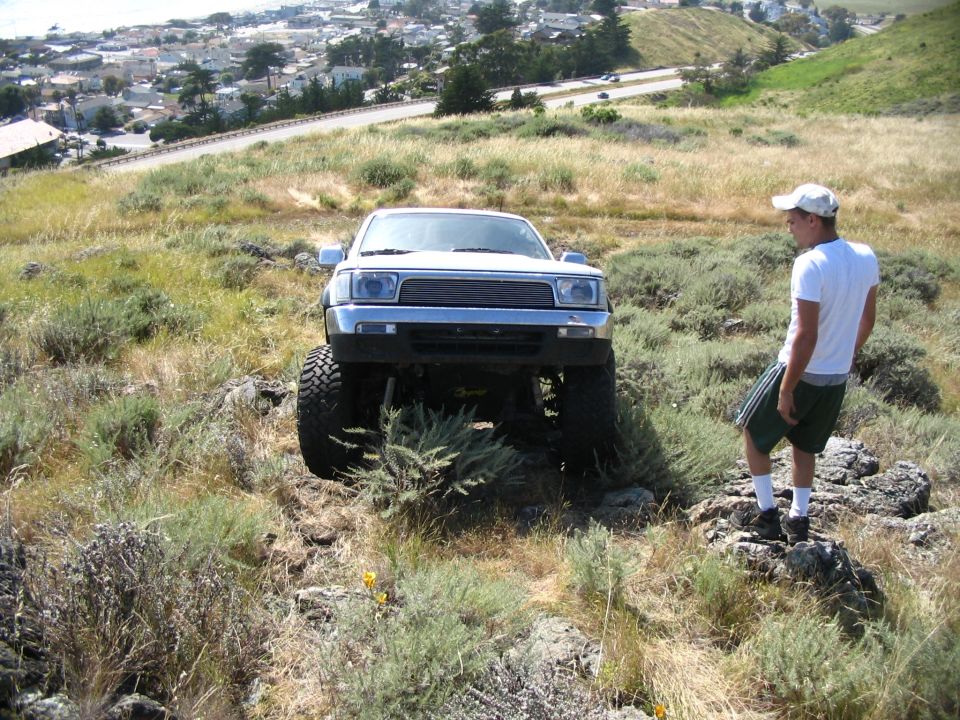 Head-on white lifted Toyota with billet grille on a hillside, man standing at right