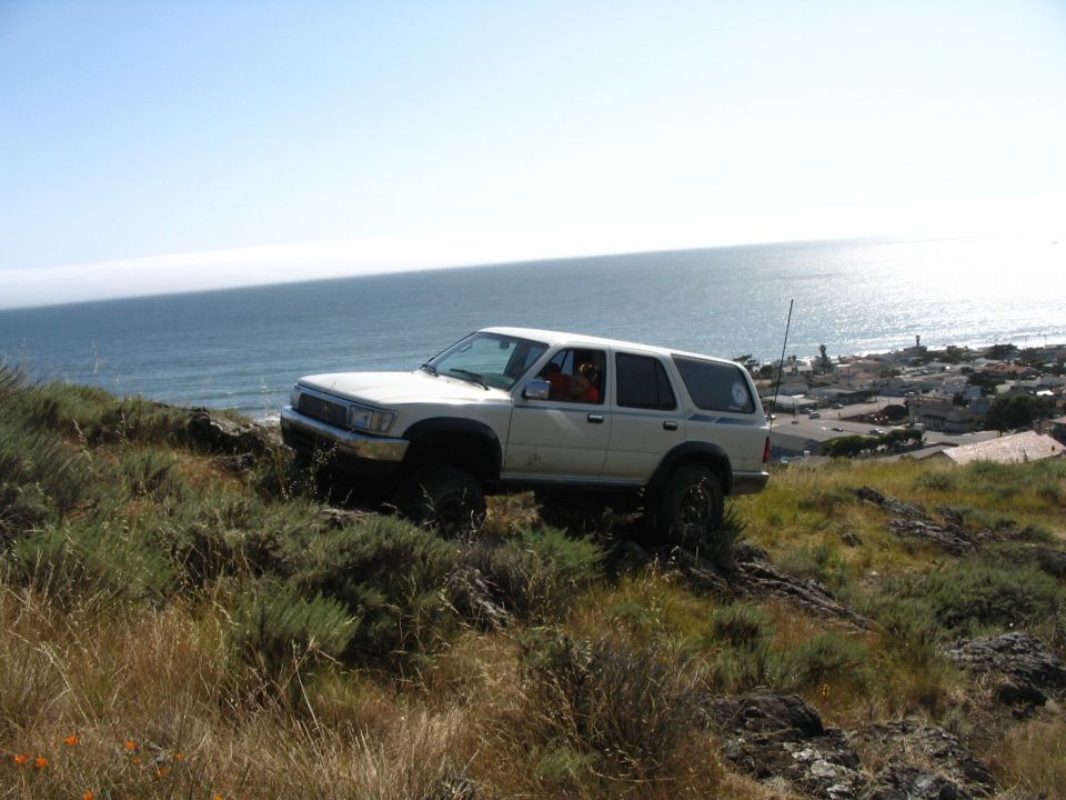 White lifted Toyota 4Runner descending a brushy coastal slope toward a sunlit ocean