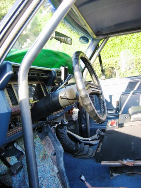 1985 Ford Bronco - The interior of a vehicle shows a roll cage, aftermarket steering wheel, and floor shifter, with blue interior panels and carpet.