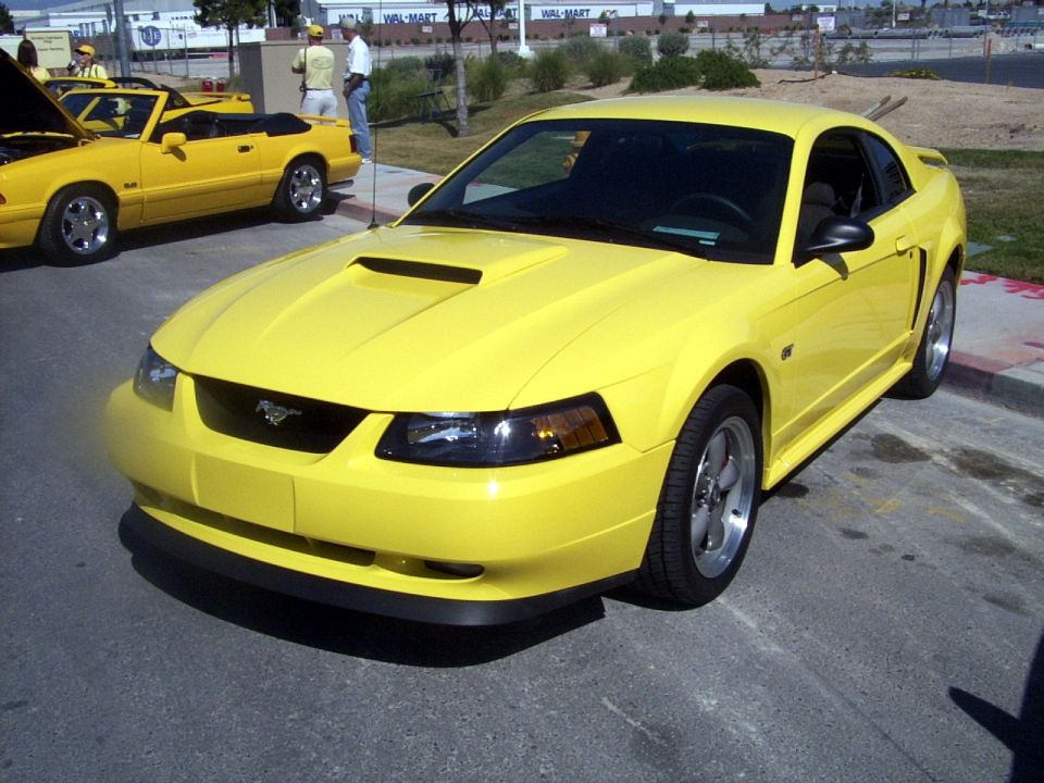 Yellow 2002 Ford Mustang parked on asphalt, another yellow convertible Mustang in background