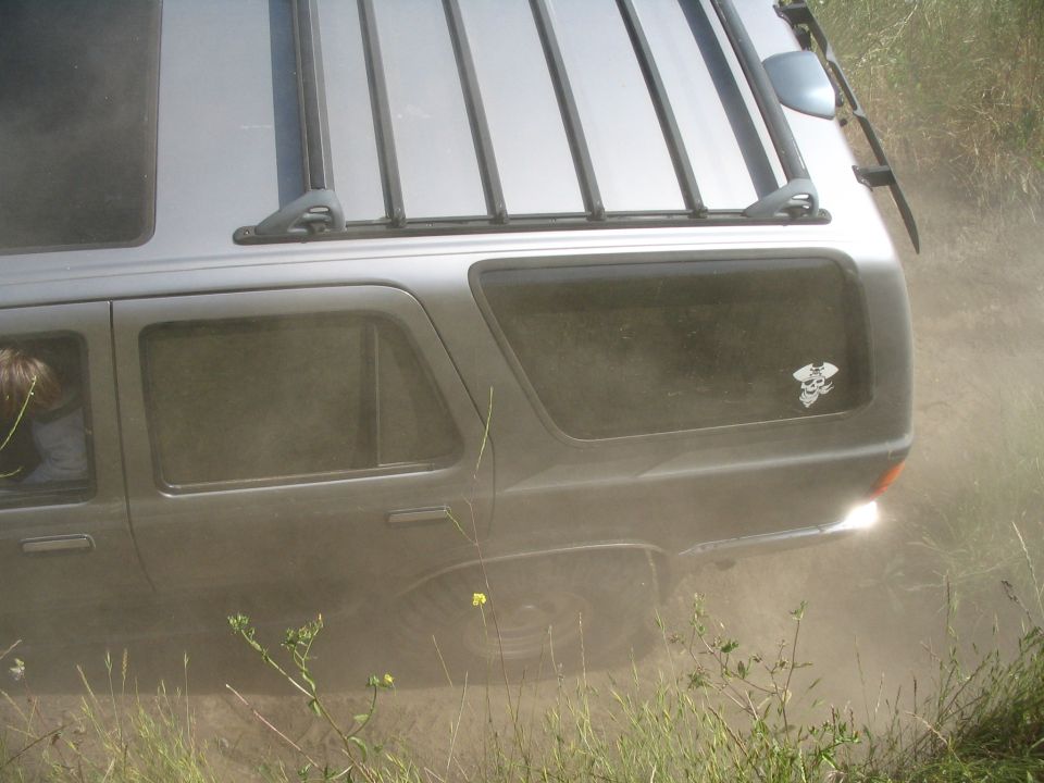 Dusty silver SUV rear quarter with roof rack and ace-of-spades decal kicking up dirt