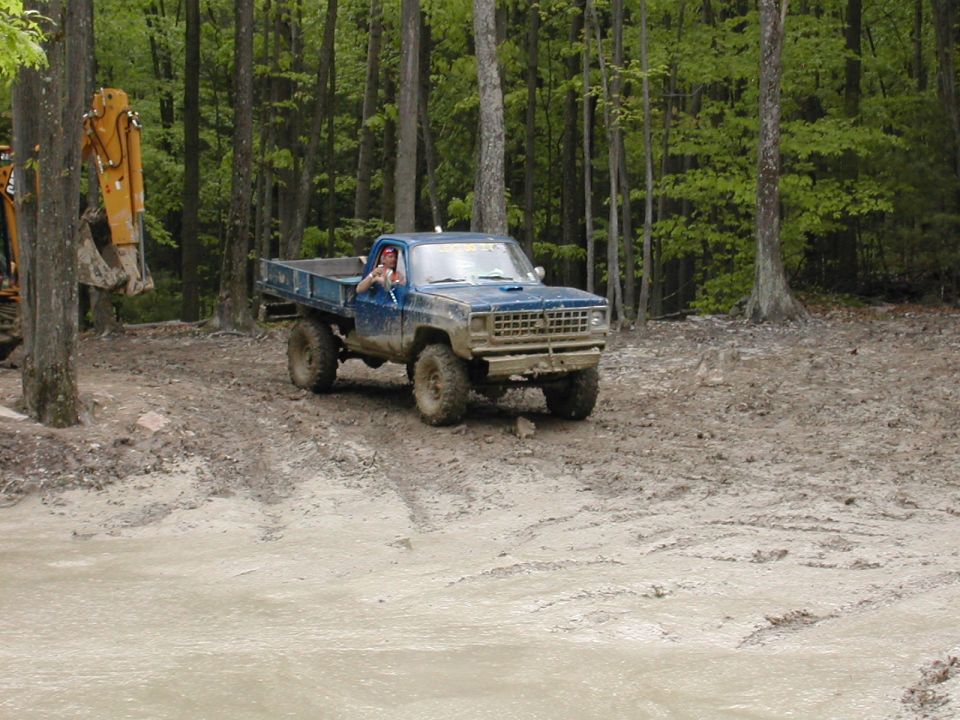 Blue pickup truck with a flatbed driving through a muddy wooded area. Driver is visible