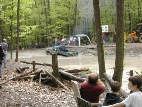 1979 Ford Bronco - A blue pickup truck is stuck in a mud pit during an event, with spectators and a backhoe nearby.