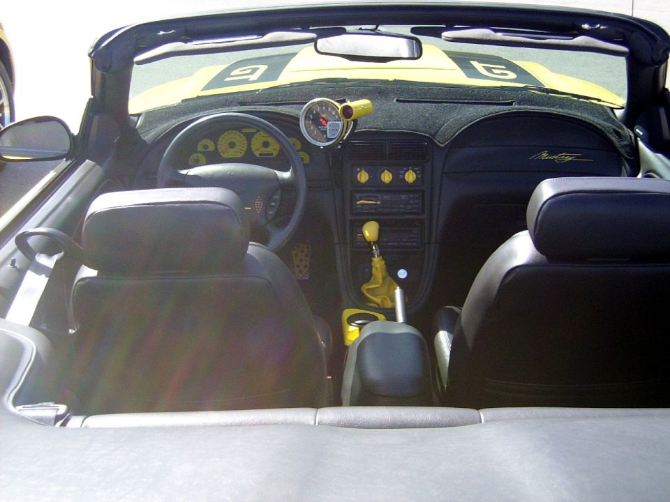 interior of a yellow convertible with black seats, aftermarket gauge on dash, and 'Mustang' script