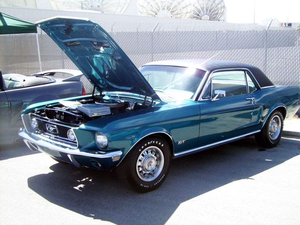 Teal 1967-1968 Ford Mustang GT coupe with hood open, showing engine bay. Another car is partially visible to the left