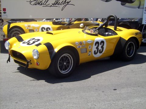 2002 Ford Mustang - Two yellow vintage race cars are parked side-by-side, with a trailer bearing Carroll Shelby's signature in the background.