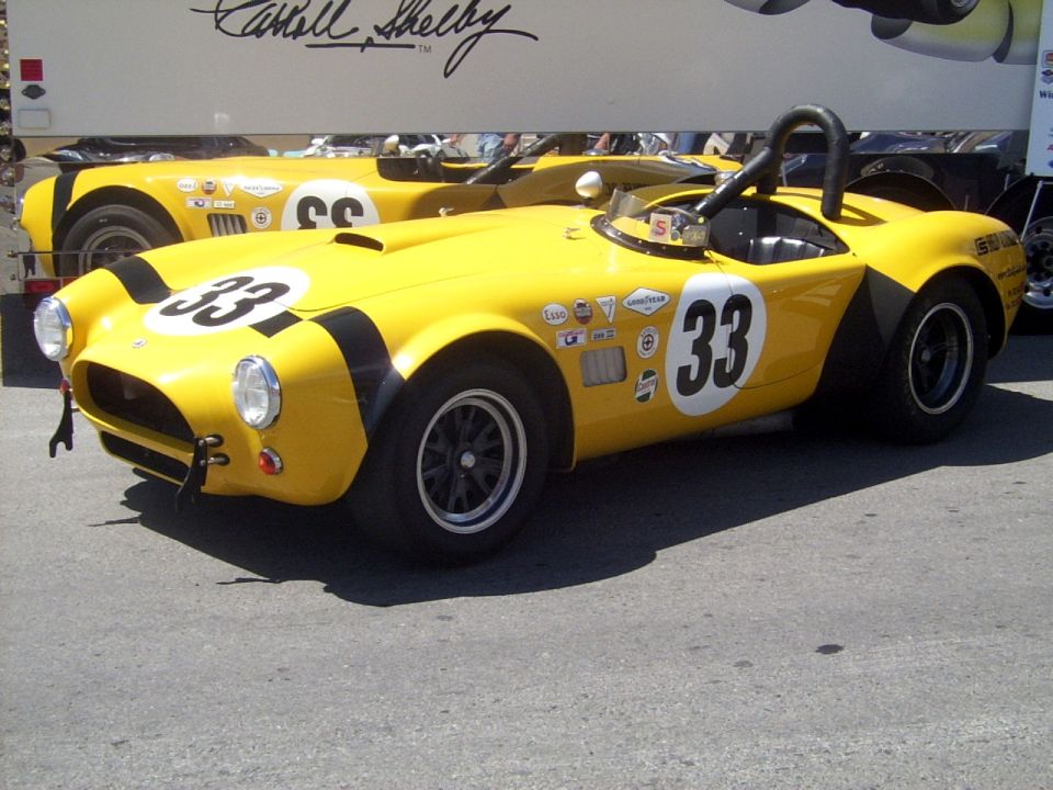 Yellow race car with black stripes and number 33 on the side. Another similar car is behind it. A trailer with Carroll Shelby's signature is visible