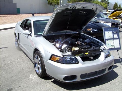 2002 Ford Mustang - A silver 2002 Ford Mustang is displayed with its hood open, revealing the engine and an informational board. Another Mustang is visible in…