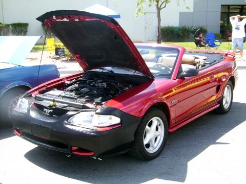2002 Ford Mustang - A red convertible with its hood open, revealing the engine bay and tan interior, parked next to a blue vehicle.