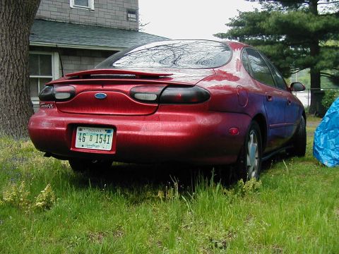 1997 Ford Taurus - Rear view of a red sedan with a spoiler and a dented rear bumper, parked in grass next to a house.