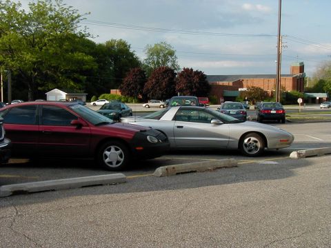 1997 Ford Taurus - A silver sports car is parked in a lot next to a red sedan, with a building and trees in the background.