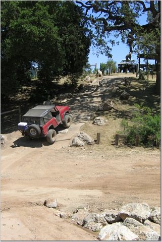 1971 Ford Bronco - A red off-road vehicle navigates a rocky dirt trail under a large tree, with another vehicle visible on a hill in the distance.