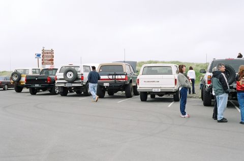 1979 Ford Bronco - Multiple SUVs and trucks are parked in a lot near a beach with a signpost in the background.