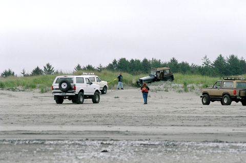 1979 Ford Bronco - Multiple off-road vehicles are gathered on a sandy terrain near a treeline under an overcast sky.