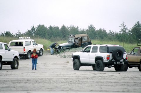 1979 Ford Bronco - Several off-road vehicles are gathered on a sandy beach with dunes and trees in the background.
