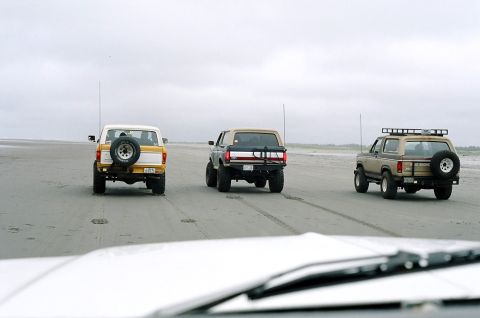 1979 Ford Bronco - Three Ford Broncos drive in formation on a beach, kicking up sand.