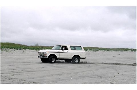 1979 Ford Bronco - A white Ford Bronco kicks up sand as it drives along a beach under a cloudy sky.