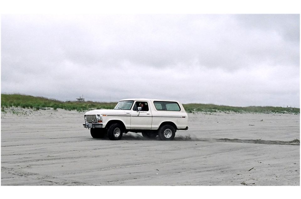 White Ford Bronco driving on a sandy beach with dunes and overcast sky